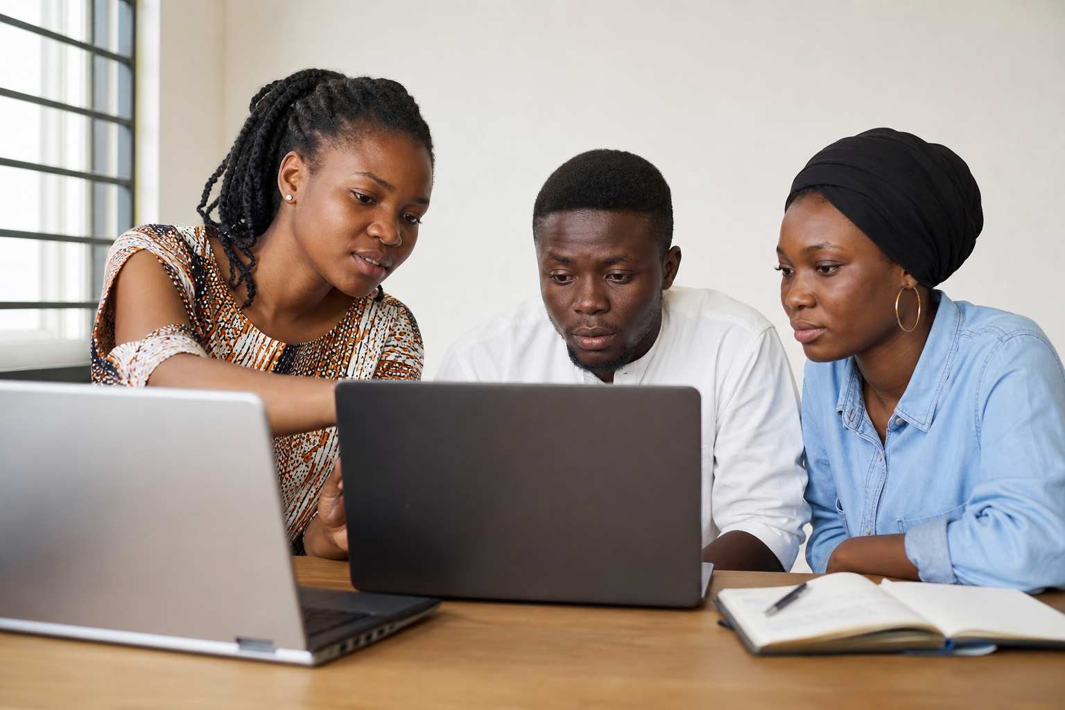 Three Nigerian young adults learning together around two laptops and a notebook in a digital work class.